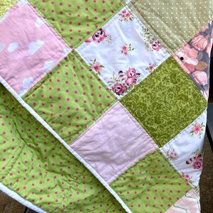 May include: A patchwork quilt with a variety of patterns and colors. The quilt features squares of green with pink polka dots, pink with white clouds, white with pink floral designs, and green floral patterns. The quilt is edged with white fabric.