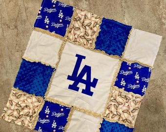 Puede incluir: Un edredón de retazos con el logotipo de los Dodgers de Los Ángeles en el centro. El edredón está hecho de varias telas, incluyendo azul, blanco y patrones de béisbol. Los bordes están adornados con un volante decorativo.