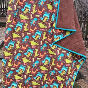 May include: A brown and turquoise dinosaur themed quilt with a repeating pattern of dinosaurs and bones. The quilt is folded in half and has a brown backing.