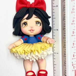 May include: A handcrafted figurine of a character with black hair, a red bow, and a blue top with red polka dots. The figurine has a yellow and white skirt and red shoes. The figurine is approximately 3 inches tall.