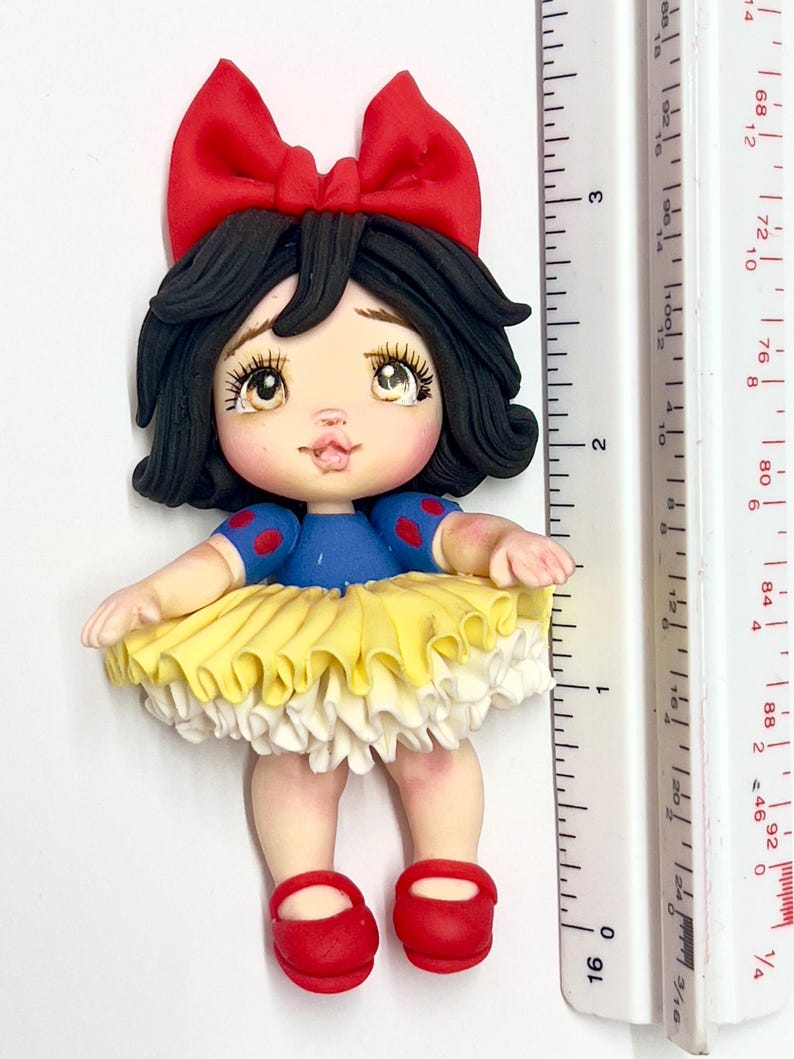 May include: A handcrafted figurine of a character with black hair, a red bow, and a blue top with red polka dots. The figurine has a yellow and white skirt and red shoes. The figurine is approximately 3 inches tall.