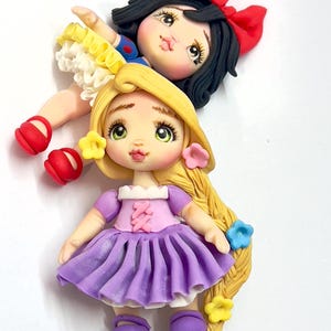 May include: Handmade polymer clay figurines of two fairytale princesses. One doll has black hair with a red bow and a blue and yellow dress. The other doll has long blonde hair, a purple dress, and flower accents.