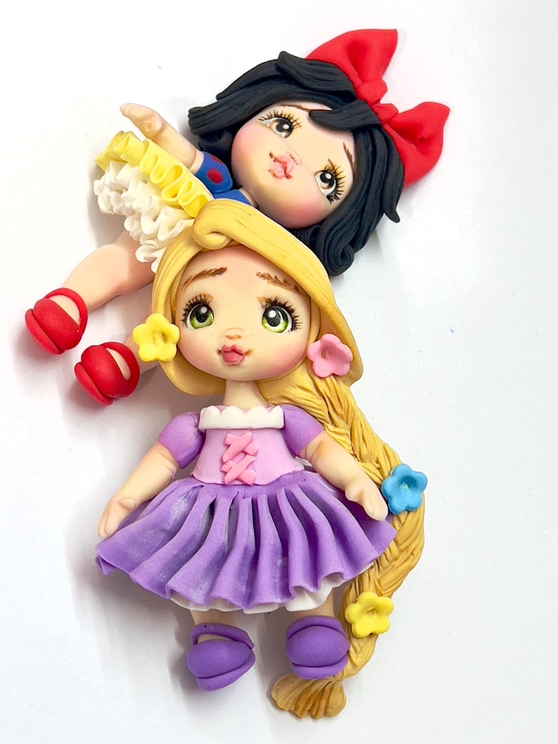 May include: Handmade polymer clay figurines of two fairytale princesses. One doll has black hair with a red bow and a blue and yellow dress. The other doll has long blonde hair, a purple dress, and flower accents.