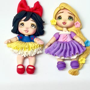May include: Two handmade figurines of cartoon characters. One is Snow White, with a red bow, black hair, and a blue and yellow dress. The other is Rapunzel, with long blonde hair, a purple dress, and flower accents. Both have detailed facial features.
