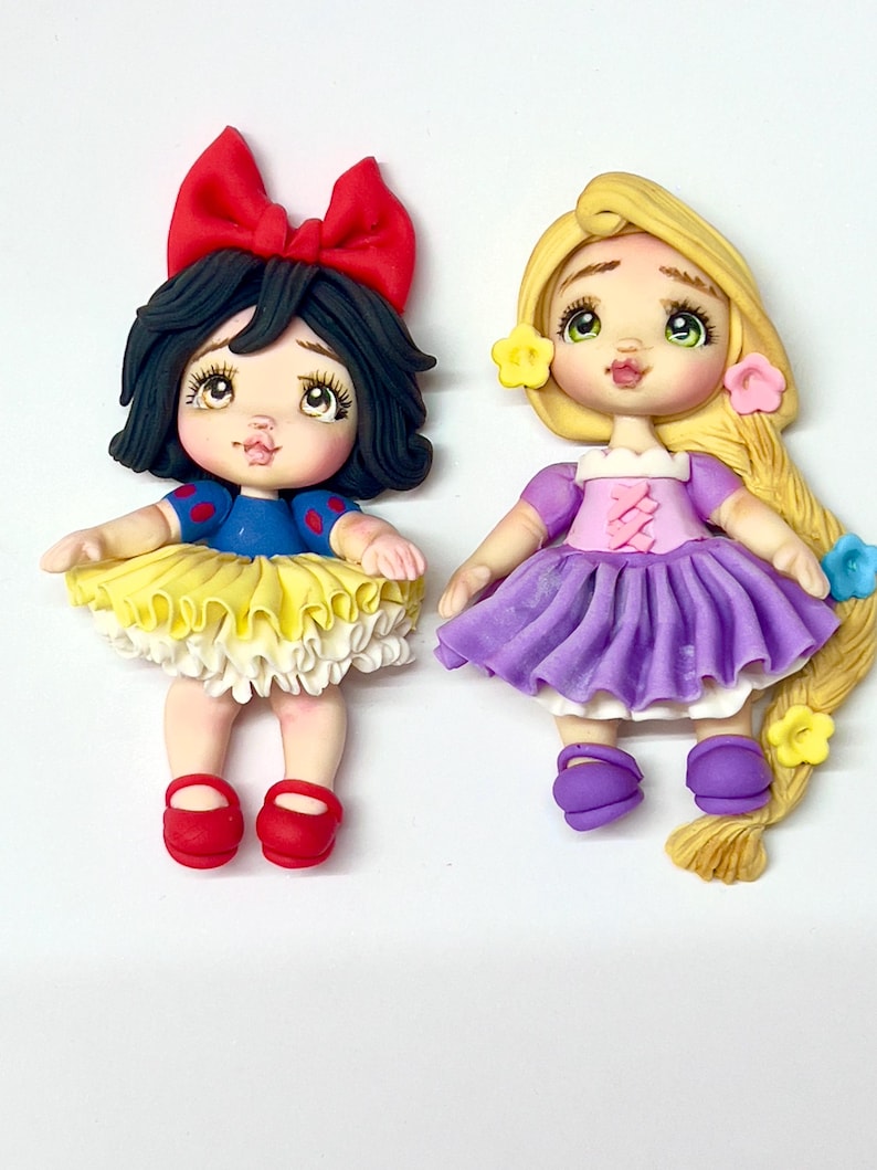 May include: Two handmade figurines of cartoon characters. One is Snow White, with a red bow, black hair, and a blue and yellow dress. The other is Rapunzel, with long blonde hair, a purple dress, and flower accents. Both have detailed facial features.