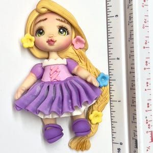 May include: A handcrafted polymer clay figurine of a princess with long blonde hair, green eyes, and a purple dress. The figurine is adorned with yellow, pink, and blue flower accents. The figure is approximately 3 inches tall.