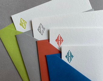 May include: Five white stationery note cards with different colored monograms in a diamond shape. The monograms are "WKB", "ECM", "MHA", "KHM". The cards are stacked on top of each other with the top card showing the "KHM" monogram. The cards are on top of four different colored envelopes: green, gray, red, and blue.