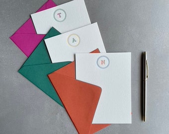 May include: Four white envelopes with different colored envelopes behind them. The envelopes have a circle with a letter in the center. The letters are T, A, and N. A silver pen is on the right side of the image.