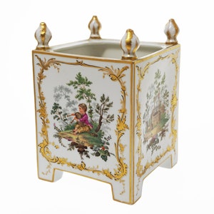 May include: A white porcelain planter with gold trim and a pastoral scene painted on each side. The planter has four legs and a square shape.