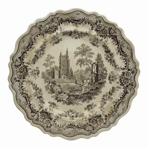 May include: A white ceramic plate with a brown and white floral pattern and a central image of a castle in a landscape. The plate has a scalloped edge.