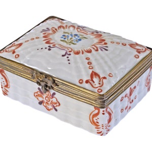 May include: A rectangular white ceramic trinket box with a brass-colored metal frame and clasp. The box is decorated with red, purple, and blue floral patterns. The box measures approximately 4 inches long, 3 inches wide, and 2 inches high.