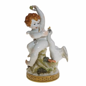 Op de afbeelding: Een porseleinen beeldje dat een cherubijn met rood haar afbeeldt, zittend op een rots, in interactie met een witte zwaan. De basis is versierd met een gouden geometrisch patroon. De cherubijn houdt een groen voorwerp vast.