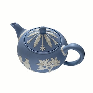 May include: A matte light blue teapot featuring white relief ornamentation. The lid displays a radial leaf pattern with a central knob, while the body is decorated with white figures and tree motifs in a classic bas-relief style.