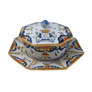 May include: A decorative ceramic serving dish with a lid, featuring a blue, yellow, and orange floral design. The dish has an octagonal shape and includes a matching octagonal plate. The lid has a blue knob and a crest design.