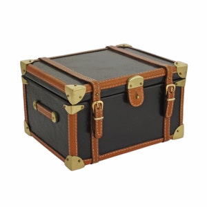 May include: A small, black trunk with tan leather trim and brass hardware. The trunk features a hinged lid, corner protectors, and leather straps with buckles. The handle is also made of tan leather. The trunk is a decorative storage box.