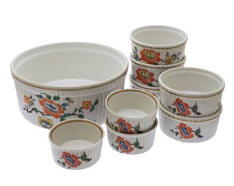 May include: A collection of cream-colored ceramic serving bowls and ramekins. The bowls and ramekins have a floral design in orange, blue, and yellow. The rims are trimmed with a gold-colored band. The ramekins have a ribbed exterior.