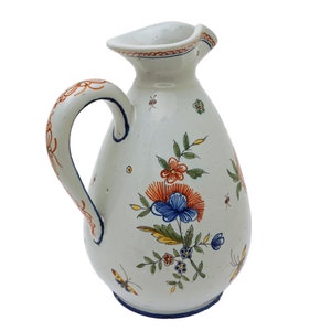 May include: White ceramic pitcher with a flared rim and a curved handle. The pitcher is decorated with colorful floral and butterfly motifs in red, blue, and orange. The rim and handle are accented with orange and blue details.