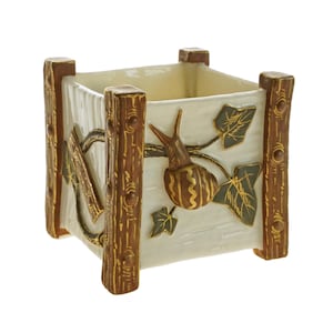 May include: A white ceramic planter with a square shape and brown wooden accents. The planter features a raised design of a snail crawling on a vine with green leaves.