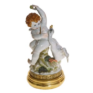 May include: A porcelain figurine of a cherub with red hair and wings, playing with a swan. The figurine is mainly white with gold detailing on the base, which features a Greek key pattern. The cherub holds a small green object.