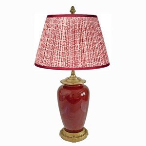 May include: A red ceramic table lamp with a patterned lampshade. The lampshade is pleated with a red and white design. The lamp has a brass base and accents. The lamp is a decorative lighting fixture.