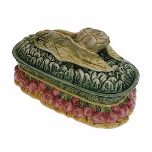May include: A ceramic vegetable-themed serving dish with a lid. The lid is green and textured, with an artichoke and leaves on top. The base is decorated with red radishes and a yellow border.