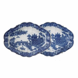 May include: Two blue and white porcelain platters with a scalloped edge. Each platter features a detailed landscape scene with a bridge, buildings, and trees. The platters are mounted with metal hangers.