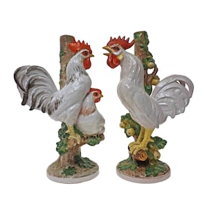 May include: Two ceramic rooster figurines. The roosters are white with red combs and yellow feet, perched on tree branches with green leaves. One figurine has a hen. The base is brown.