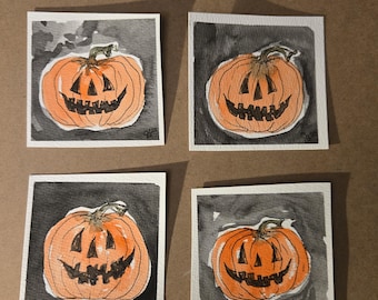 May include: Four watercolor paintings of orange jack-o'-lanterns with black, smiling faces and green stems. Each pumpkin is set against a dark gray and black background within a white square frame. The artwork is on a brown surface.