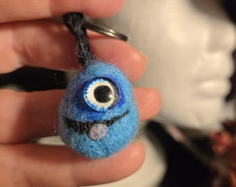 May include: A small, round, blue needle-felted creature keychain with a single large, realistic eye featuring a blue iris and black pupil. The creature has a stitched black mouth and a small, light purple tongue, attached to a metal keyring.