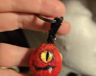 May include: A round, red needle-felted keychain featuring a single large, yellow, reptilian-style eye with a vertical black pupil. The creature has a stitched black mouth with a single white tooth and a black pipe cleaner attached to a metal ring.