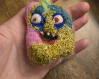 May include: A needle-felted creature ornament featuring a pink side, a textured olive green side, and a blue face area. It has two white eyes with red pupils, a wide black mouth with visible white teeth, and a tuft of light green hair on top.