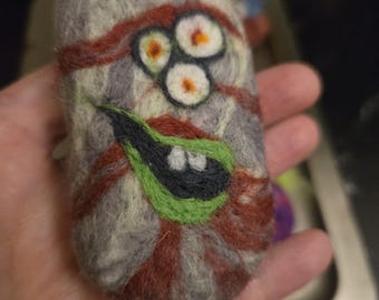 May include: A rectangular felted wool object featuring a mottled gray and brown base with swirling patterns. The surface displays three circular white eyes with orange centers and a wide, black mouth outlined in bright green with two white teeth.