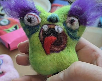 May include: A needle-felted creature figure in bright lime green with prominent purple tufts of hair on top. The figure features two wide, bulging eyes, a dark open mouth with visible white teeth, and a protruding red tongue, all crafted from wool.