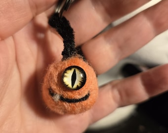 May include: A round, orange needle-felted keychain charm featuring a single large, yellow eye with a vertical black pupil. The creature has a small, stitched black mouth with a tiny white tooth and a black fuzzy stem extending from the top.