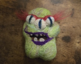 May include: A needle-felted monster figure in a light green color with three white eyes featuring dark blue pupils. The creature has a wide purple mouth with visible white teeth, red fiber hair on the sides, and a textured, fuzzy surface.