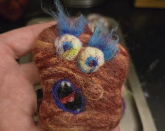 May include: A needle-felted creature made of brown wool with a textured, lumpy surface. It features two large, round eyes with blue pupils and wild, wispy blue hair on top. The creature has an open, dark mouth with a small red tongue visible inside.