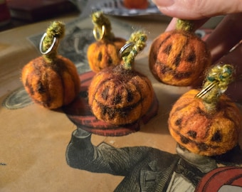 May include: Five small, round, orange needle-felted pumpkins with dark brown stitched facial features. Each pumpkin has a textured green stem with a silver-toned metal jump ring attached to the top for hanging, creating a set of miniature ornaments.