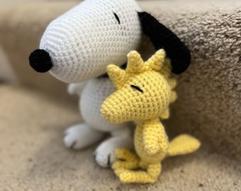 May include: A white crocheted Snoopy plush toy with black eyes and a black nose, hugging a yellow crocheted Woodstock plush toy with black eyes.