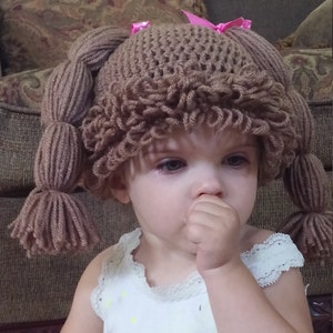 May include: A child wearing a brown crocheted hat with pigtails and pink bows. The hat is designed to look like a doll's wig.