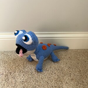 May include: A blue crocheted lizard toy with orange spots and large white eyes. The toy has a long tail and is lying on a light brown carpet.