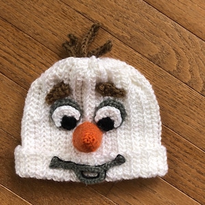 May include: White crocheted hat with a snowman face. The hat has brown yarn for hair, black eyes, an orange nose, and a gray mouth.