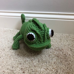 May include: A green crocheted chameleon toy with black and white eyes. The toy is lying on a beige carpet.
