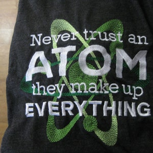 May include: A dark gray t-shirt with white embroidery that reads "Never trust an ATOM they make up EVERYTHING". A green embroidered atom symbol is behind the text.