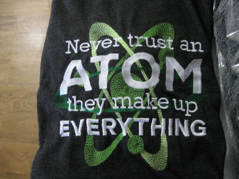 May include: A dark gray t-shirt with white embroidery that reads "Never trust an ATOM they make up EVERYTHING". A green embroidered atom symbol is behind the text.