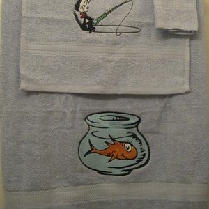 May include: A set of three blue bath towels with embroidered images of a cat wearing a hat fishing for a fish in a bowl. The towels have a white border.