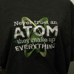 May include: A dark grey t-shirt with a white embroidered design. The design features a green atom symbol and the text "Never trust an ATOM they make up EVERYTHING".