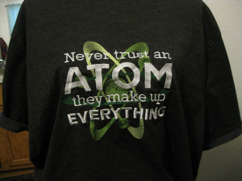 May include: A dark grey t-shirt with a white embroidered design. The design features a green atom symbol and the text "Never trust an ATOM they make up EVERYTHING".