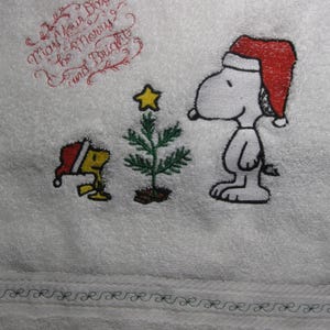 May include: Embroidered white towel with a Christmas theme featuring Snoopy and Woodstock. The text reads "May Your Days Be Merry and Bright".