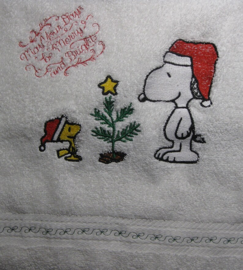 May include: Embroidered white towel with a Christmas theme featuring Snoopy and Woodstock. The text reads "May Your Days Be Merry and Bright".
