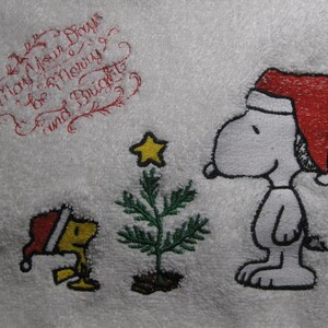 May include: Embroidered design featuring Snoopy wearing a red Santa hat, standing next to a small Christmas tree with a yellow star on top. A small yellow bird with a red Santa hat is to the left of the tree. The text "May Your Days Be Merry & Bright" is embroidered in red cursive at the top of the image.